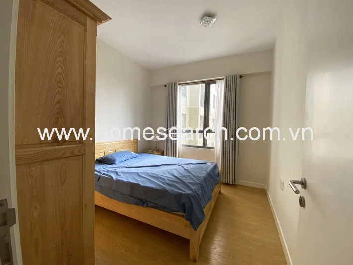 This bedroom features warm wood-tone flooring and a matching wooden bed frame and wardrobe, creating a cohesive, natural aesthetic. A large window with neutral-toned curtains provides ample natural light and a view of the adjacent building, while a wall-mounted AC unit ensures climate control.