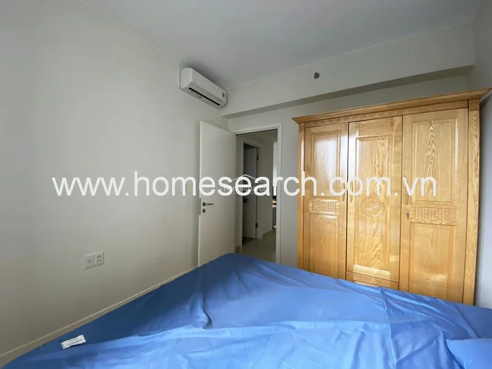 The bedroom layout is functional, with the wooden wardrobe positioned conveniently near the entrance and a wall-mounted AC unit above the doorway. The neutral wall color and wood flooring provide a clean, minimalist backdrop that maximizes the sense of space in the room.