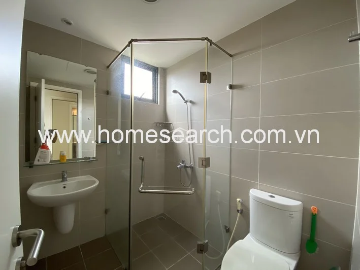 This bathroom is finished with neutral-toned wall and floor tiles, featuring a modern glass-enclosed walk-in shower and a wall-mounted pedestal sink. The space is well-maintained and includes a large vanity mirror, a bidet sprayer, and a small window for ventilation.