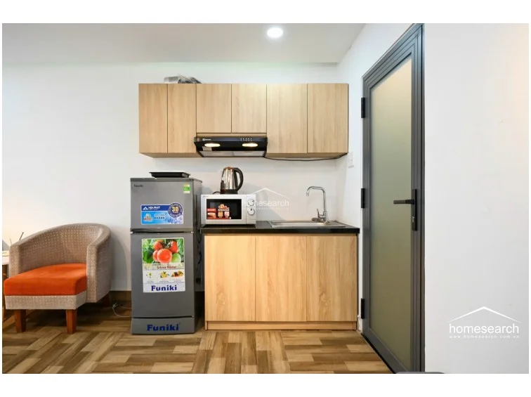 This compact kitchenette features light wood-grain cabinetry, a black countertop, and a stainless steel sink, providing efficient storage and preparation space. It is equipped with a Funiki refrigerator, a microwave, and a range hood, all set against clean white walls and durable wood-look tile flooring.