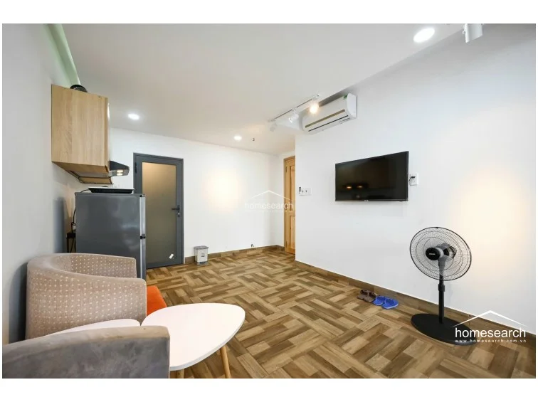 This open-plan living area integrates a kitchenette with a cozy seating nook, featuring a modern armchair and a small coffee table. The space is well-maintained with white walls, wood-look tile flooring, and a wall-mounted television, complemented by a portable fan and efficient recessed lighting.