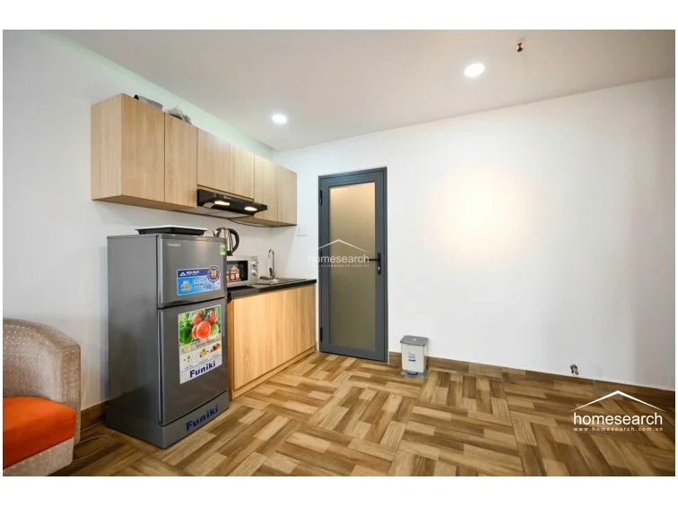 This view of the kitchenette highlights the efficient use of space with light wood cabinets and a stainless steel sink, positioned conveniently next to a frosted glass door. The area maintains a minimalist aesthetic with white walls and wood-look tile flooring, presenting a clean and well-kept environment.