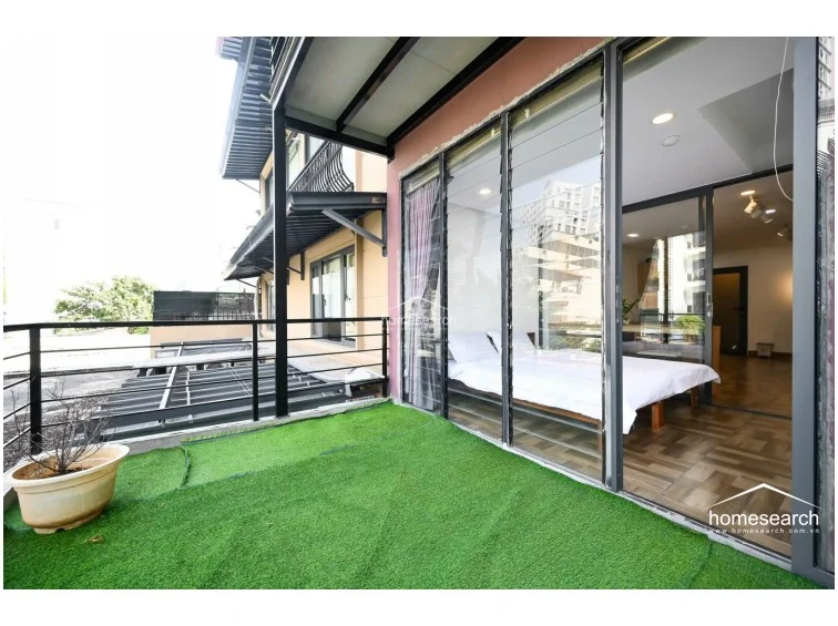 The unit includes a private, expansive outdoor terrace covered in artificial turf, providing a versatile space for relaxation or outdoor dining. This balcony connects directly to the bedroom through large, floor-to-ceiling glass sliding doors, which allow for an abundance of natural light and an open, airy flow. The area is enclosed by a black metal railing and offers an urban view, enhancing the overall appeal of the property.