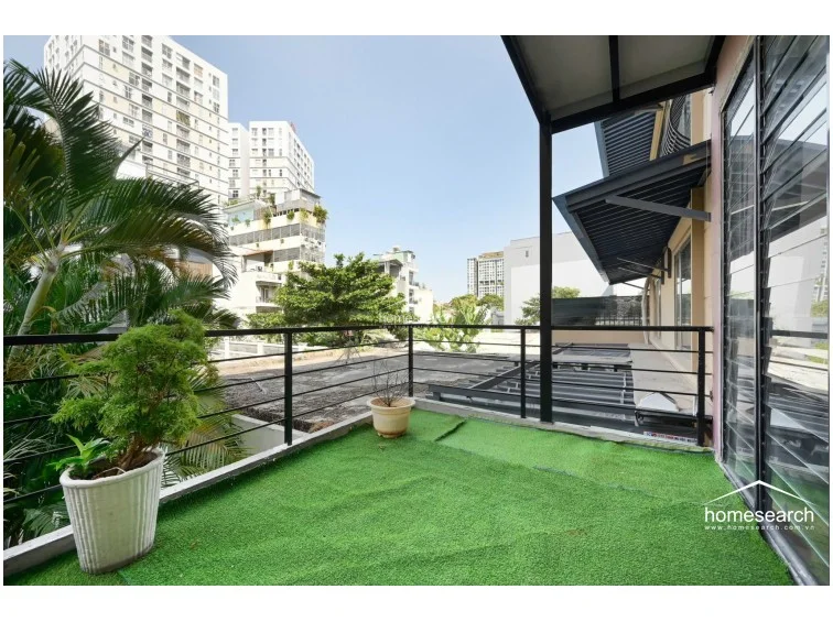 This wide-angle view of the terrace highlights its generous size and direct connection to the interior living space via large glass panels. The area is equipped with a black metal safety railing and features a potted plant, adding a touch of greenery to the urban setting. The terrace overlooks a neighboring building complex, offering a clear view of the surrounding city landscape.