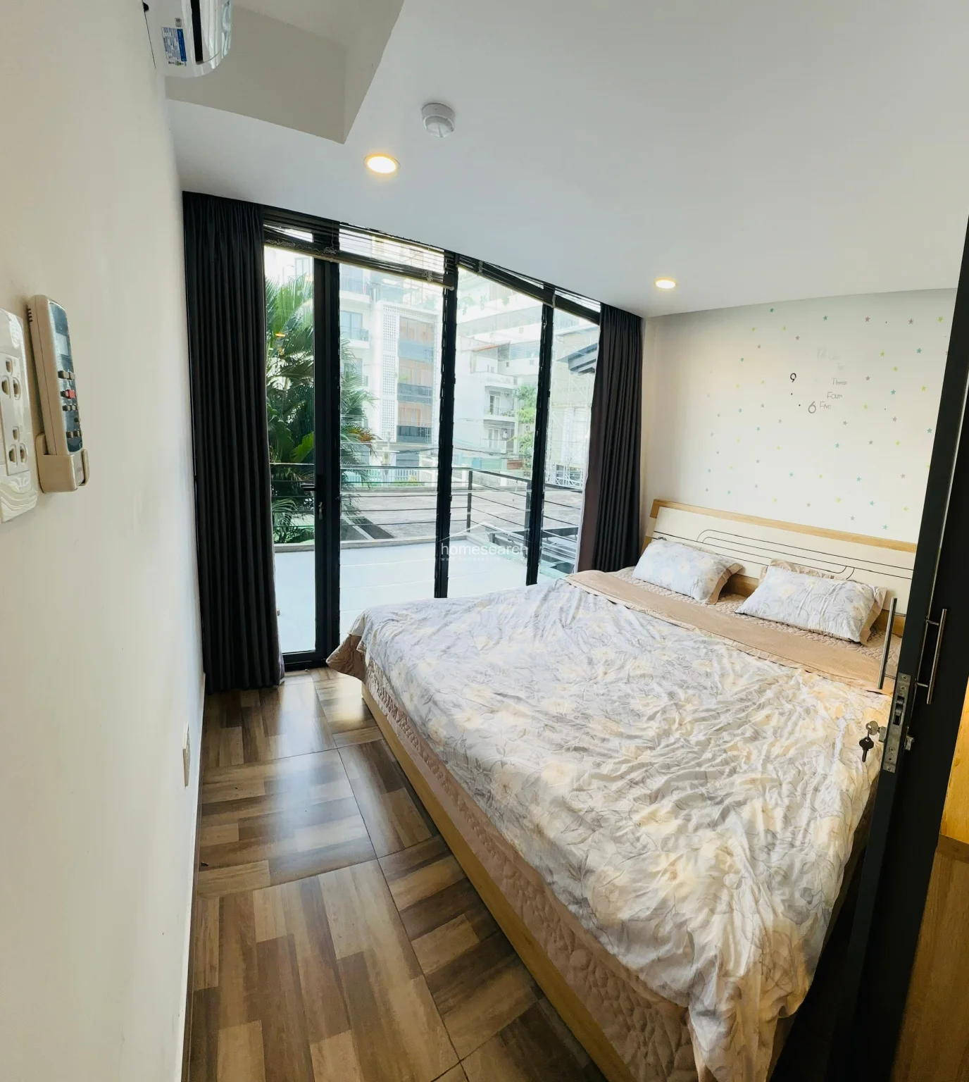 This bright bedroom features a comfortable double bed with patterned linens and is bathed in natural light from the expansive floor-to-ceiling glass doors that lead to a private balcony. The room is finished with wood-look tile flooring and white walls adorned with playful star decals, creating a modern and airy atmosphere.