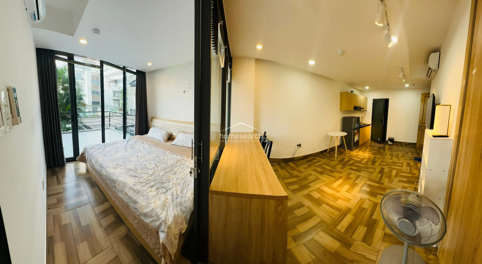 This panoramic view shows a well-organized studio layout that seamlessly connects a bedroom with a glass partition to a functional living and kitchenette area. The space feels spacious and bright, highlighted by the wood-look tile floors, light wood cabinetry, and a private balcony that offers a pleasant outdoor connection.