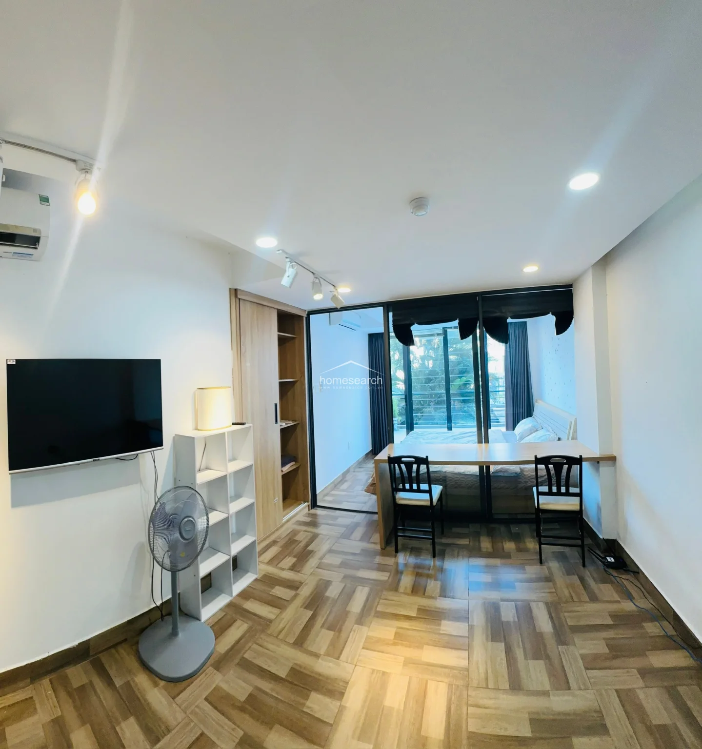 This studio apartment features a modern, open-concept layout with a distinct separation between the living/workspace and the sleeping area provided by a glass partition. The space is finished with wood-look tile flooring, white walls, and track lighting, creating a bright and contemporary atmosphere. Essential amenities include a wall-mounted AC unit, a flat-screen TV, a built-in wooden wardrobe, and a functional desk setup that overlooks the bedroom area.