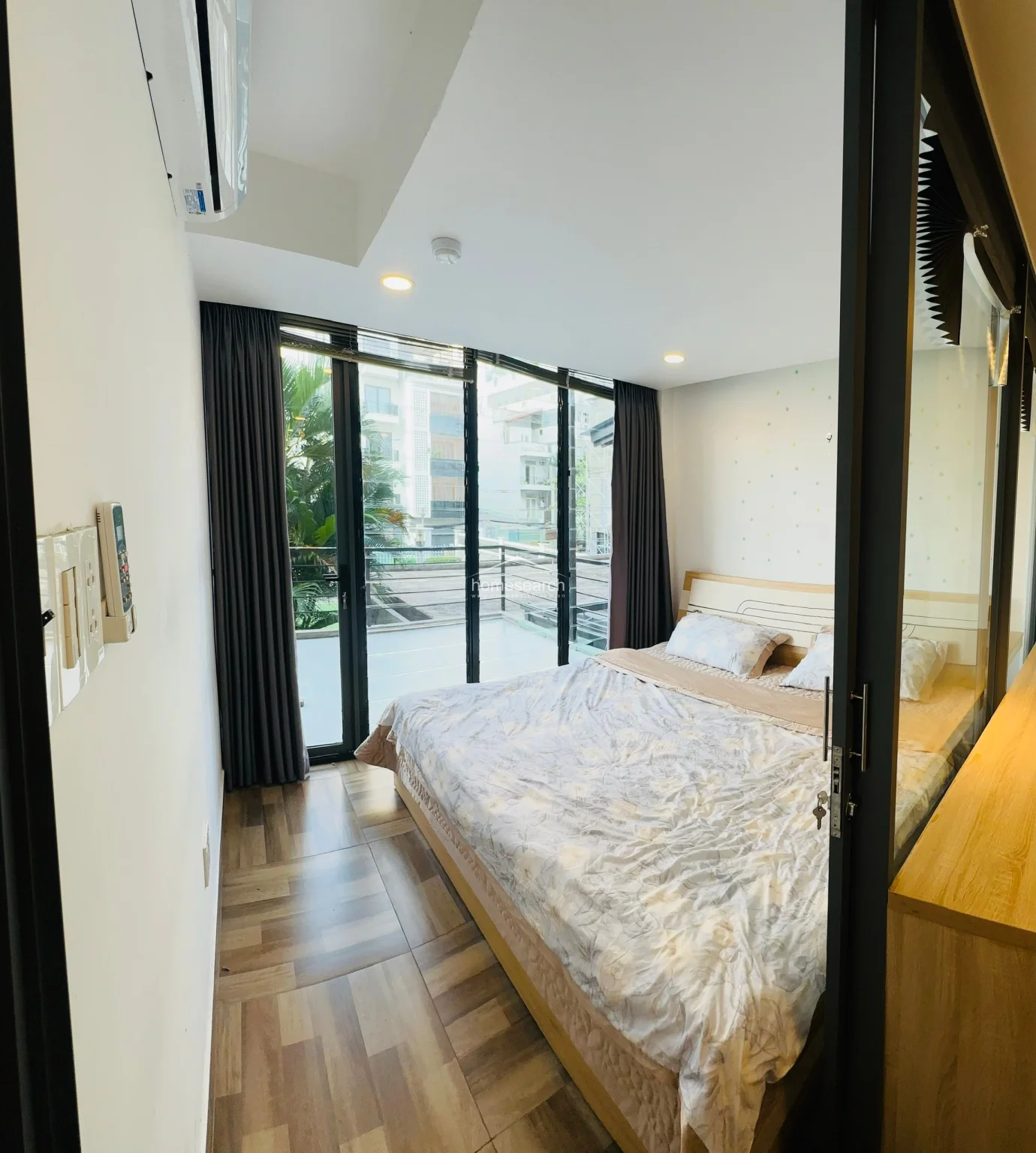 The bedroom is designed as a cozy, private retreat, separated from the main living area by a sleek glass wall that maintains a sense of spaciousness. It features a comfortable bed with neutral bedding, dark curtains for privacy, and direct access to the outdoor terrace. The room maintains a consistent aesthetic with the rest of the unit, utilizing the same wood-patterned flooring and recessed ceiling lighting.