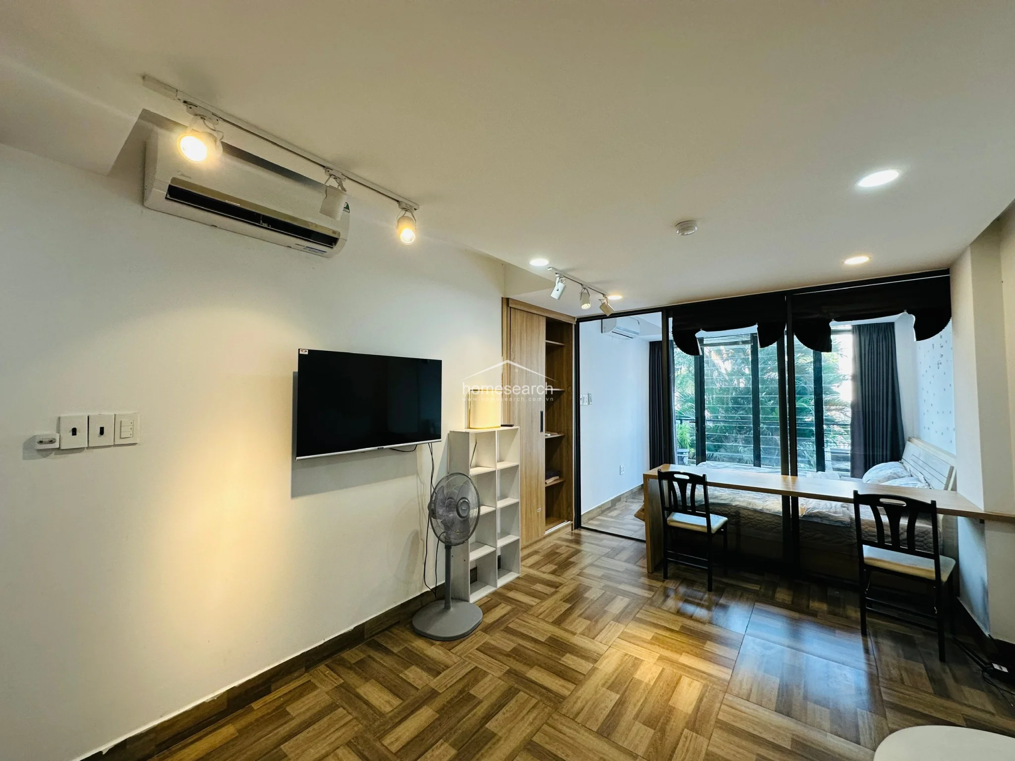 This view emphasizes the functional layout of the studio, showcasing the seamless transition between the living area and the bedroom. The space is well-maintained and efficiently furnished with a white shelving unit, a desk, and a wall-mounted television, making it ideal for a minimalist lifestyle. The combination of track lighting and natural light from the terrace ensures the unit remains bright and welcoming throughout the day.