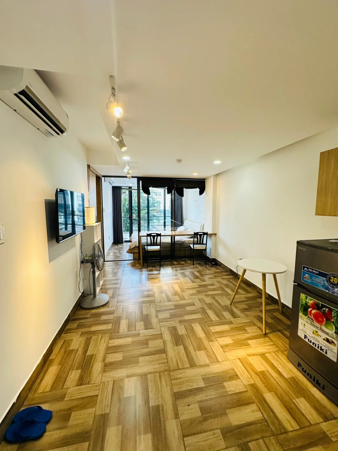 This studio apartment features an open-concept layout with a seamless flow from the kitchenette area to the sleeping zone, separated by a glass partition. The space is brightened by a large floor-to-ceiling window with a balcony view, complemented by light-toned wood-look tile flooring and crisp white walls. It is well-equipped with a wall-mounted AC unit, track lighting, a flat-screen TV, and a compact kitchenette with light wood cabinetry.