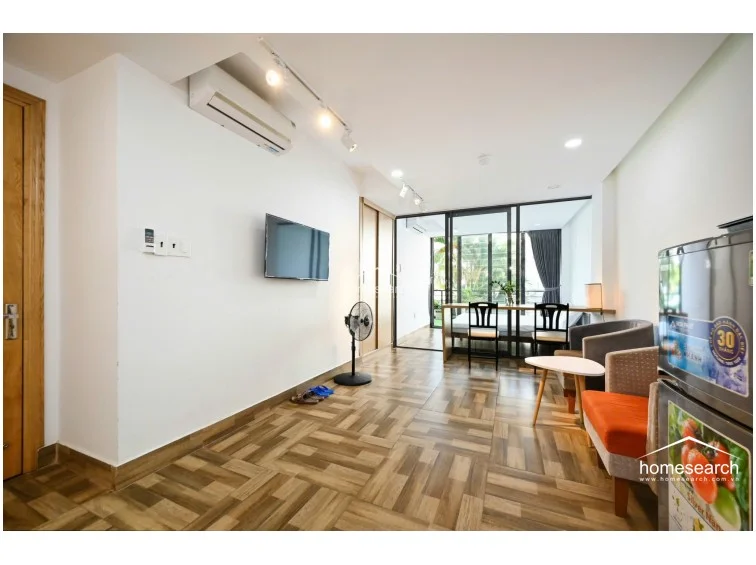 This open-plan studio apartment features a modern, functional layout with wood-look tile flooring and crisp white walls. The space is efficiently divided by a glass partition that separates the living area from the bedroom, allowing for natural light to flow throughout the unit while maintaining a sense of openness.