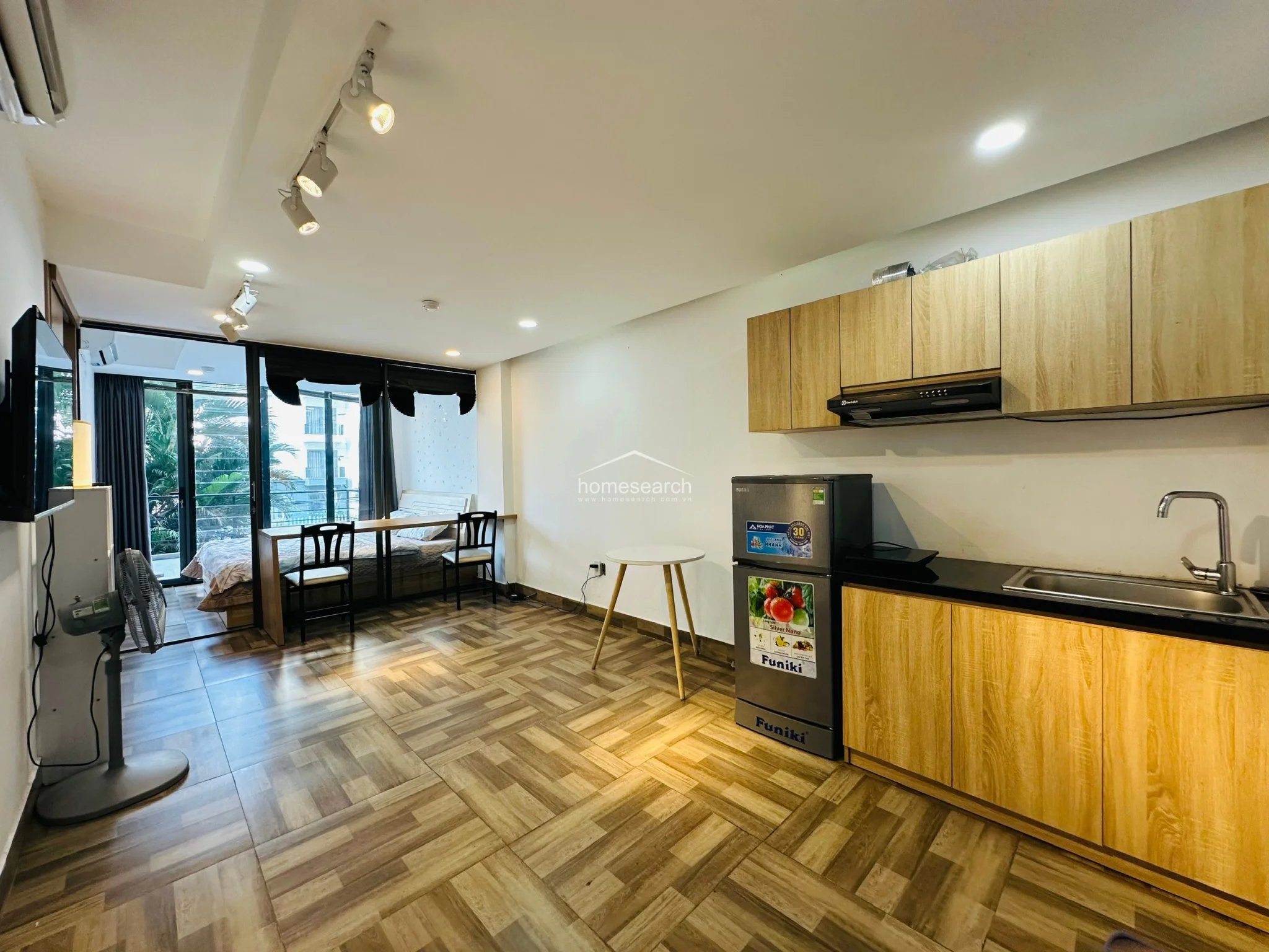 The kitchenette area is efficiently designed with light wood upper and lower cabinets, a black countertop, and a built-in exhaust hood above the cooking area. A modern, compact refrigerator is included, and the overall aesthetic is clean and minimalist, maintaining a consistent color scheme with the rest of the unit. The space feels well-maintained and functional for a studio living arrangement.