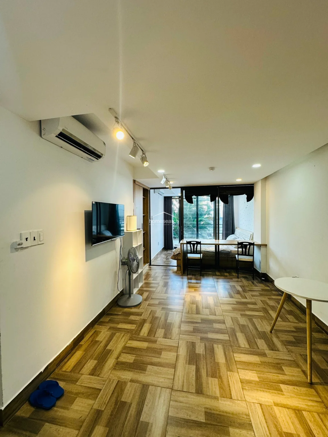 The living area is equipped with a wall-mounted television, a split-system AC unit, and a compact seating arrangement, creating a comfortable lounge space. A sleek, light-wood desk with two chairs sits against the glass partition, providing a dedicated workspace that overlooks the bedroom and the outdoor balcony beyond.