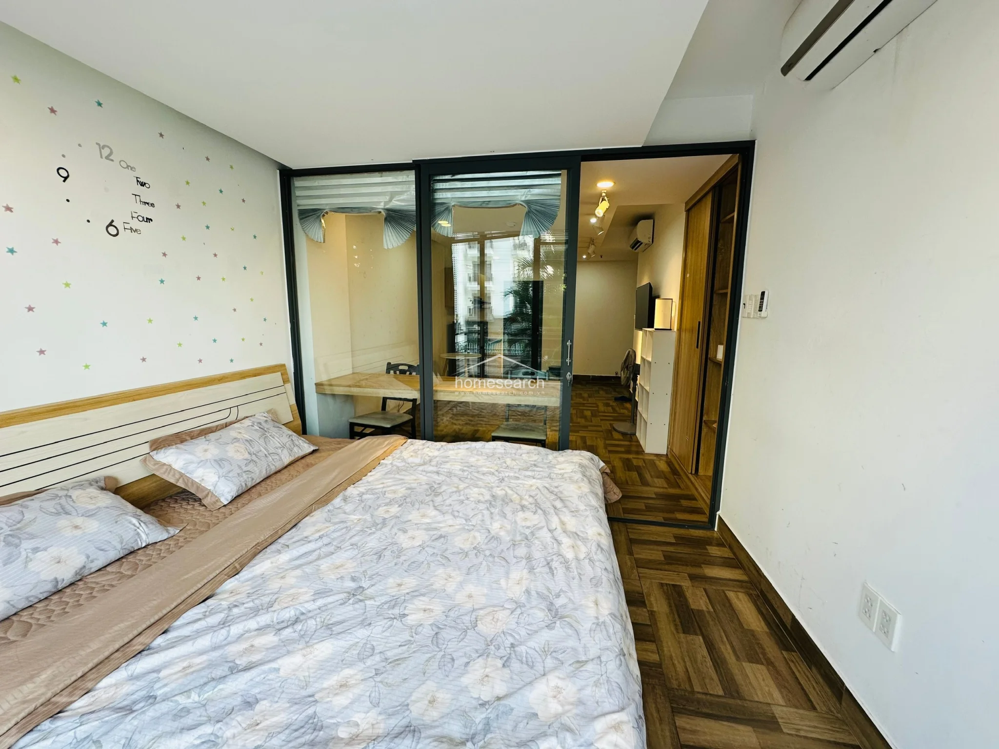 The bedroom area is partitioned by a sleek black-framed glass wall, allowing natural light to filter through from the balcony while maintaining a sense of privacy. The room features a comfortable bed with floral bedding and a playful star-patterned accent wall, creating a cozy atmosphere. The flooring continues the wood-look tile pattern, ensuring visual continuity throughout the entire unit.