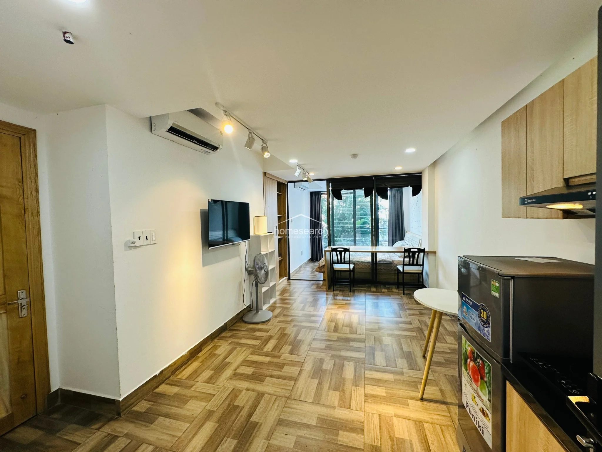 This studio offers a highly functional layout, highlighting the transition from the living/kitchen area to the bedroom through the glass sliding doors. The room is outfitted with modern amenities, including a wall-mounted television, a portable fan, and a dedicated workspace at the foot of the bed. The neutral color palette and consistent flooring materials contribute to an organized and airy feel.