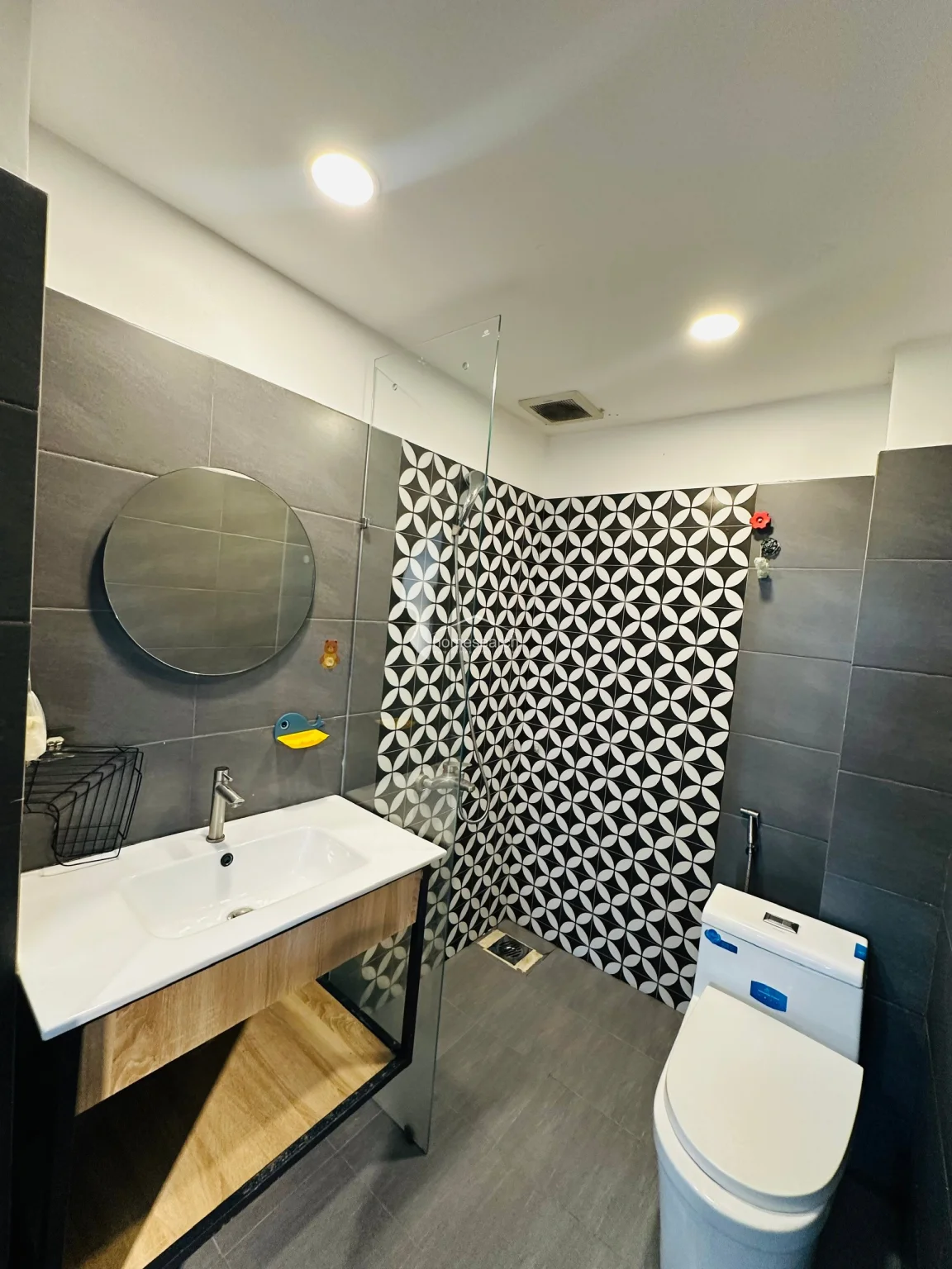 The bathroom features a contemporary design with a striking contrast between large-format grey wall tiles and a bold, black-and-white patterned accent wall in the shower area. It includes a modern vanity with a white basin, a circular mirror, and a glass shower partition for a clean, open look. The space is well-maintained and includes essential fixtures like a toilet and a handheld showerhead, presenting a stylish and updated aesthetic.