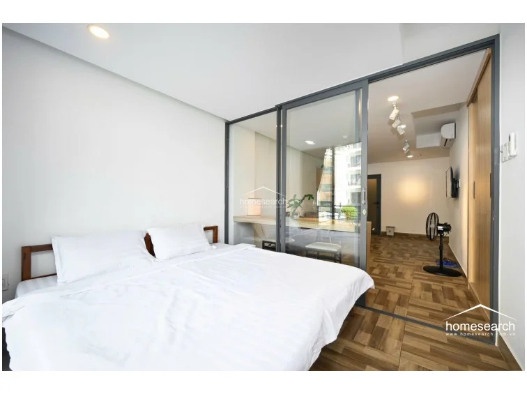 The bedroom is partitioned by floor-to-ceiling glass panels, which creates a bright, airy feel while offering privacy through dark curtains. The room includes a simple bed frame and provides direct access to a balcony, which is visible through the large window and brings in plenty of natural light.