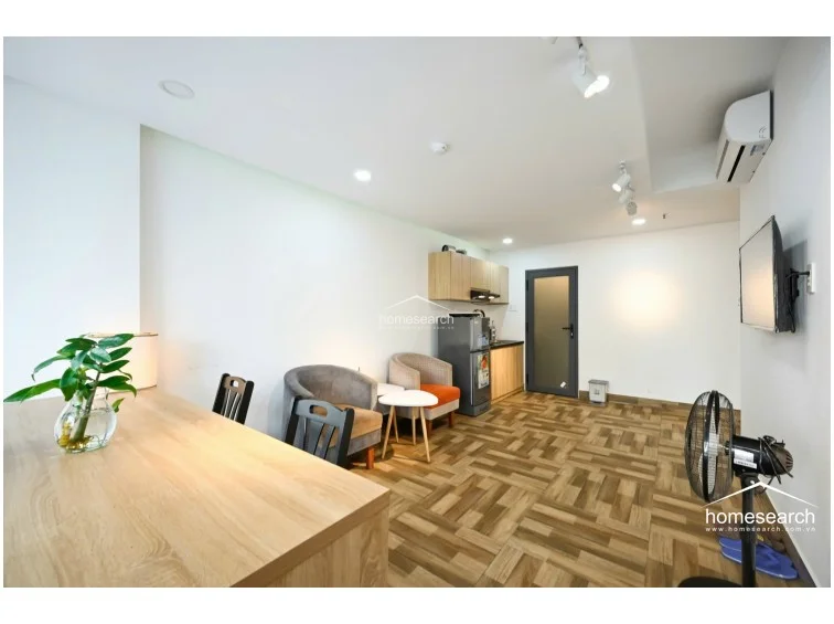 The kitchen area is compact and integrated into the main living space, featuring light-wood cabinetry and a small refrigerator. A dark-framed door leads to other parts of the property, and the overall design maintains a clean, minimalist aesthetic with recessed ceiling lighting.