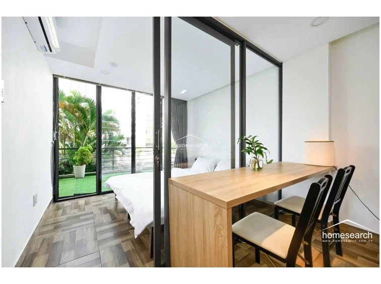 This bedroom features a sleek, modern design with a glass partition that separates it from the living area, creating an open yet defined flow. Large floor-to-ceiling windows provide abundant natural light and direct access to a private balcony with artificial turf and lush greenery. The room is finished with wood-look tile flooring and clean white walls, maintaining a bright and minimalist aesthetic.