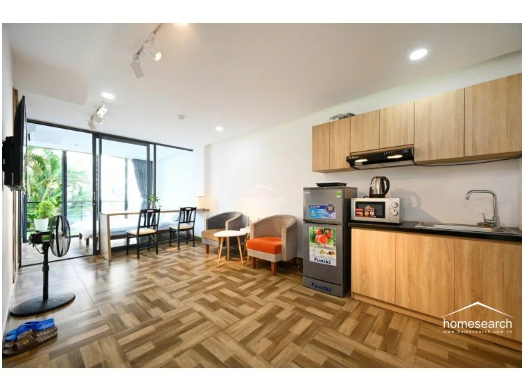 This open-plan studio layout integrates a living and dining area with a compact kitchenette, featuring light wood cabinetry and a modern black countertop. The space is efficiently furnished with a two-tone armchair set and a breakfast bar, while the wood-patterned tile flooring provides a cohesive look throughout. Large glass doors lead to a balcony, ensuring the living space remains well-lit and connected to the outdoors.