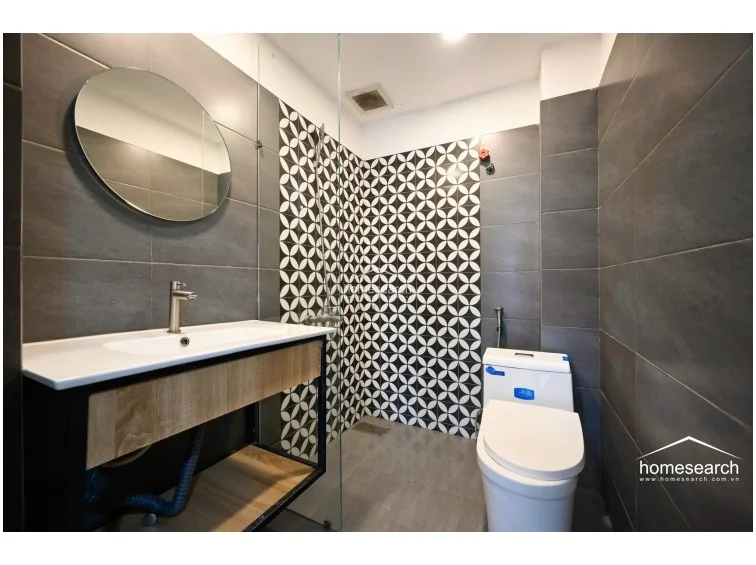 The bathroom showcases a contemporary design with a striking contrast between dark grey wall tiles and a decorative, geometric black-and-white patterned accent wall in the shower area. It is equipped with a modern vessel sink on a floating wood vanity, a circular mirror, and a glass-enclosed walk-in shower. The space is well-maintained and features a clean, minimalist aesthetic with efficient use of vertical space.