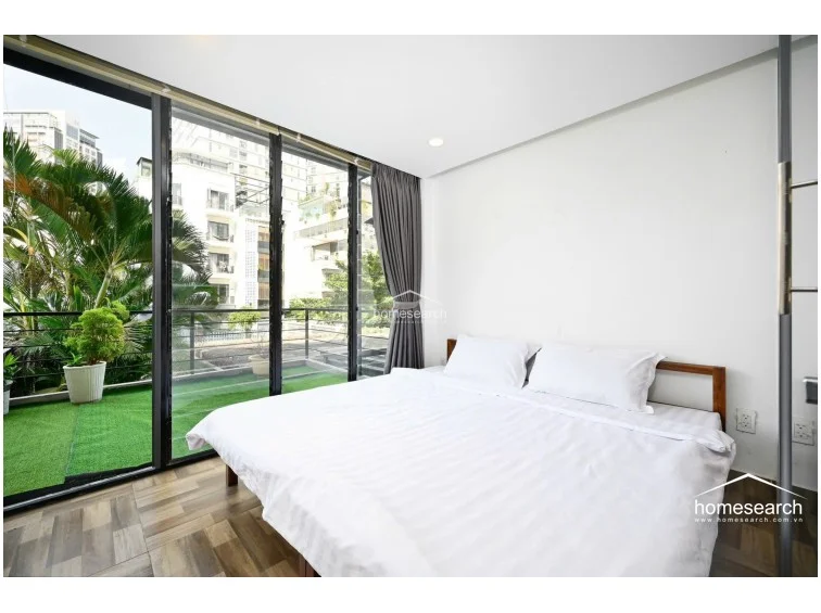This bedroom offers a serene atmosphere with a large bed positioned directly against floor-to-ceiling glass walls that overlook a private balcony. The space is bathed in natural light and features neutral white walls and wood-look tile flooring, creating a calm and airy environment. The glass partition allows for a seamless visual connection to the rest of the unit while maintaining privacy through integrated grey curtains.