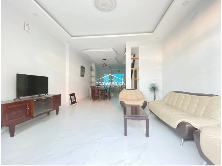 This living room features an open-concept layout that flows directly into the dining and kitchen areas, creating a spacious and airy atmosphere. The room is finished with polished white marble-effect tile flooring, crisp white walls, and a sophisticated recessed ceiling with integrated lighting. The space is furnished with a modern beige sofa set and a wooden media console, presenting a clean, well-maintained, and move-in-ready condition.
