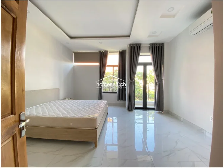 This bedroom features a clean, modern aesthetic with white walls and polished marble-look tile flooring. It includes a double bed, a wall-mounted AC unit, and direct access to a private balcony through glass doors, which provides ample natural light and an outdoor view.