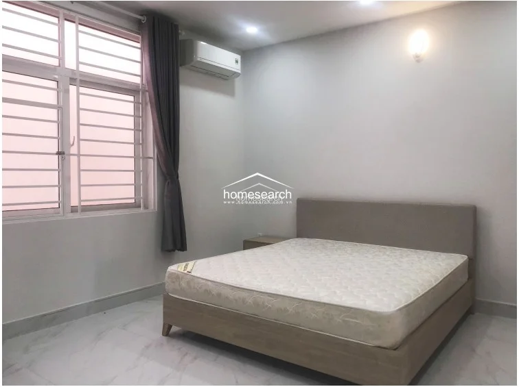 This bedroom angle emphasizes the window placement, which features security bars and grey curtains to control light and privacy. The space is finished with consistent white walls and reflective tile flooring, maintaining a cohesive and minimalist design throughout the room.