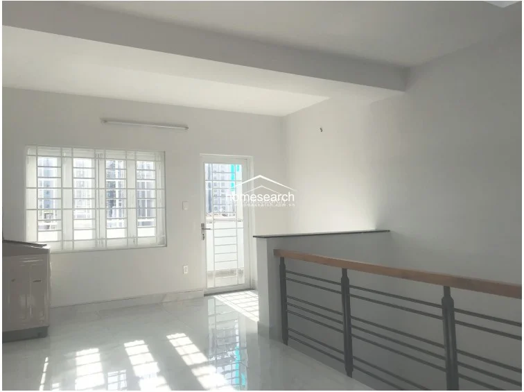 This image shows a landing or hallway area that connects to a balcony, featuring a metal railing with a wooden handrail. The space is illuminated by natural light from the glass door and window, and the polished tile floors reflect the bright, clean condition of the property.