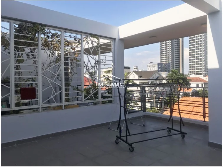 This spacious outdoor terrace offers an open-air layout with a concrete floor and a partial overhead structure. It features decorative metal security grilles with climbing greenery and provides an expansive urban view of the surrounding neighborhood and high-rise buildings.