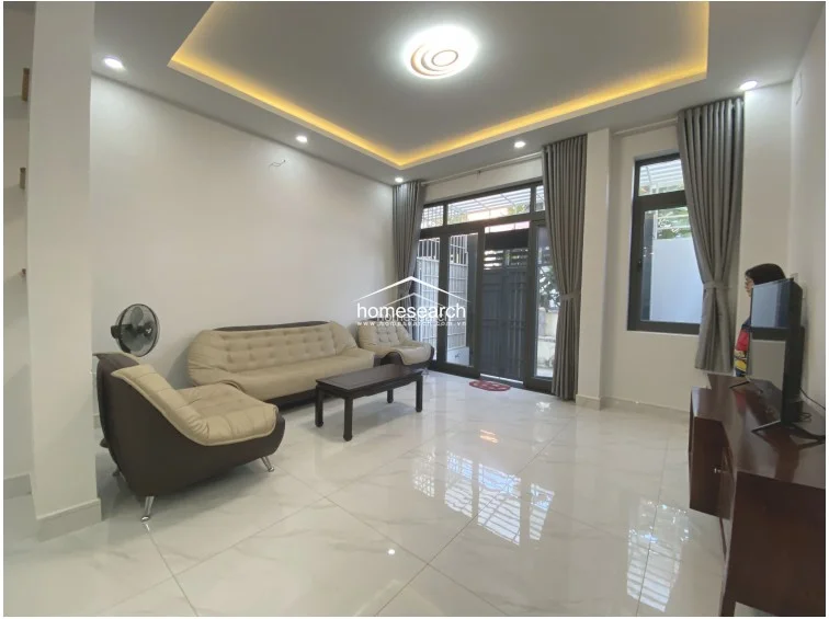 This view of the living room highlights the elegant recessed ceiling design with warm ambient cove lighting and recessed spotlights. Large windows with floor-to-ceiling grey curtains allow for significant natural light, while the white marble-effect flooring enhances the brightness of the space. The room is neatly arranged with a comfortable seating area, reflecting a high level of maintenance and a contemporary design scheme.