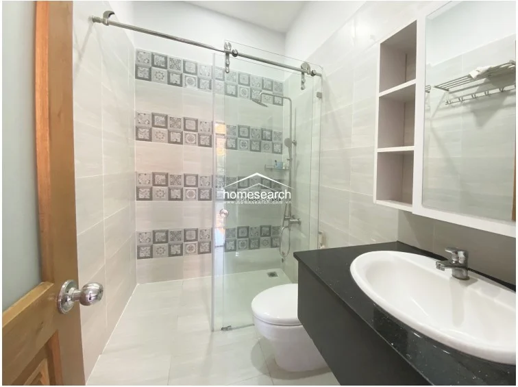 This modern bathroom features a sleek glass-enclosed walk-in shower area accented by decorative patterned wall tiles. The space is finished with light-colored floor and wall tiles, a black granite vanity countertop, and a white integrated sink, all appearing to be in excellent, well-maintained condition.