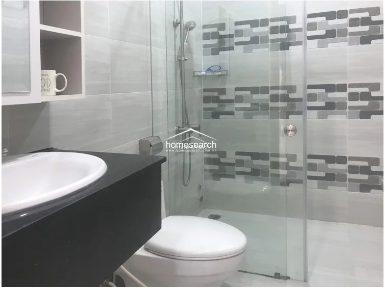 This view highlights the functional layout of the bathroom, showcasing a contemporary sliding glass shower door and a wall-mounted vanity unit with open shelving. The room utilizes a neutral color palette with geometric grey-toned accent tiles, providing a clean and organized aesthetic with efficient use of space.