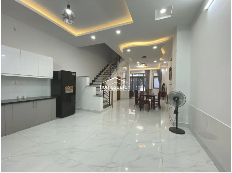 This open-plan kitchen and dining area features a modern, minimalist kitchen with white upper cabinets and grey lower cabinetry, providing a clean and functional workspace. The dining area is centrally located, flowing seamlessly from the living room and adjacent to the staircase, which features a stylish black metal railing. The space is bright and well-lit by a combination of recessed ceiling lights and a modern pendant light fixture over the dining table.