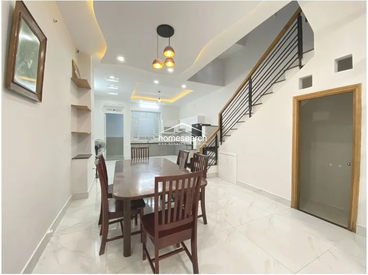 This dining area serves as the central hub of the home, connecting the kitchen and living spaces with an open flow and consistent white marble-effect tile flooring. The area is defined by a wooden dining set and a unique, curved recessed ceiling with warm accent lighting, adding architectural interest to the room. A wooden staircase with a sleek black metal railing leads to the upper floor, while the walls are kept in a neutral, pristine white finish.