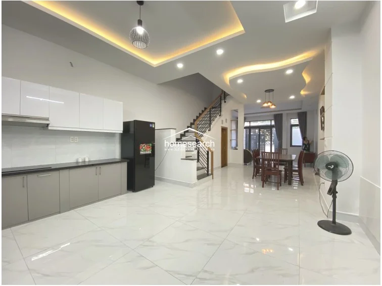 This open-concept ground floor features a modern kitchen with sleek white upper cabinets and grey lower cabinetry, seamlessly flowing into a dining and living area. The space is finished with high-gloss marble-look tile flooring, recessed cove lighting, and a central staircase with a metal railing, all maintained in excellent, move-in-ready condition.