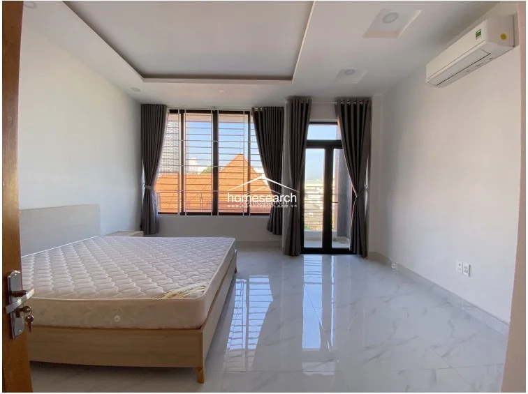 This spacious bedroom features large windows and a glass door leading to a private balcony, allowing for abundant natural light and ventilation. The room is equipped with a wall-mounted AC unit, neutral grey curtains, and polished marble-look tile flooring, creating a clean and minimalist aesthetic.