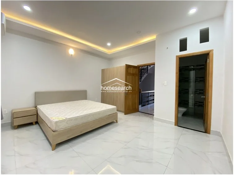 This primary bedroom includes a dedicated en-suite bathroom accessed through a wooden door, with a built-in wooden wardrobe providing ample storage. The room features a modern bed frame with a matching nightstand, recessed cove lighting, and a consistent, high-quality finish throughout.