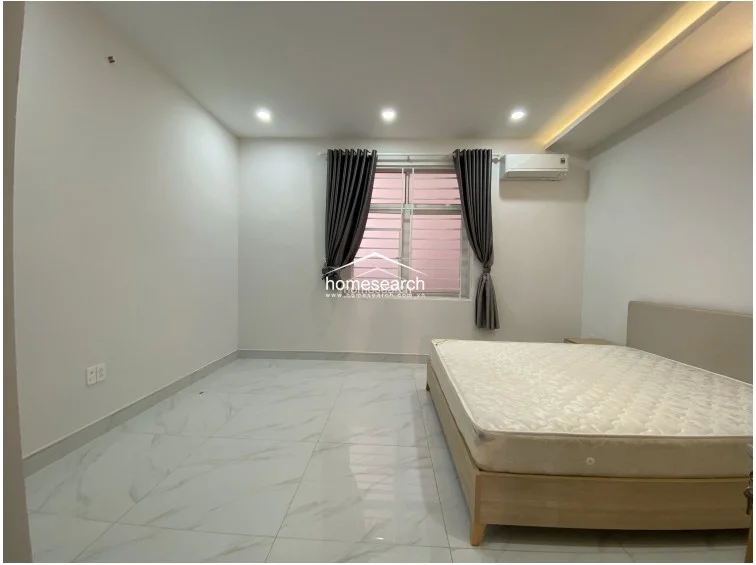 This bedroom is designed with a minimalist approach, featuring a neutral color palette and polished tile flooring that enhances the sense of space. It is equipped with a wall-mounted AC unit and a large window with grey curtains, ensuring a comfortable and well-ventilated environment.