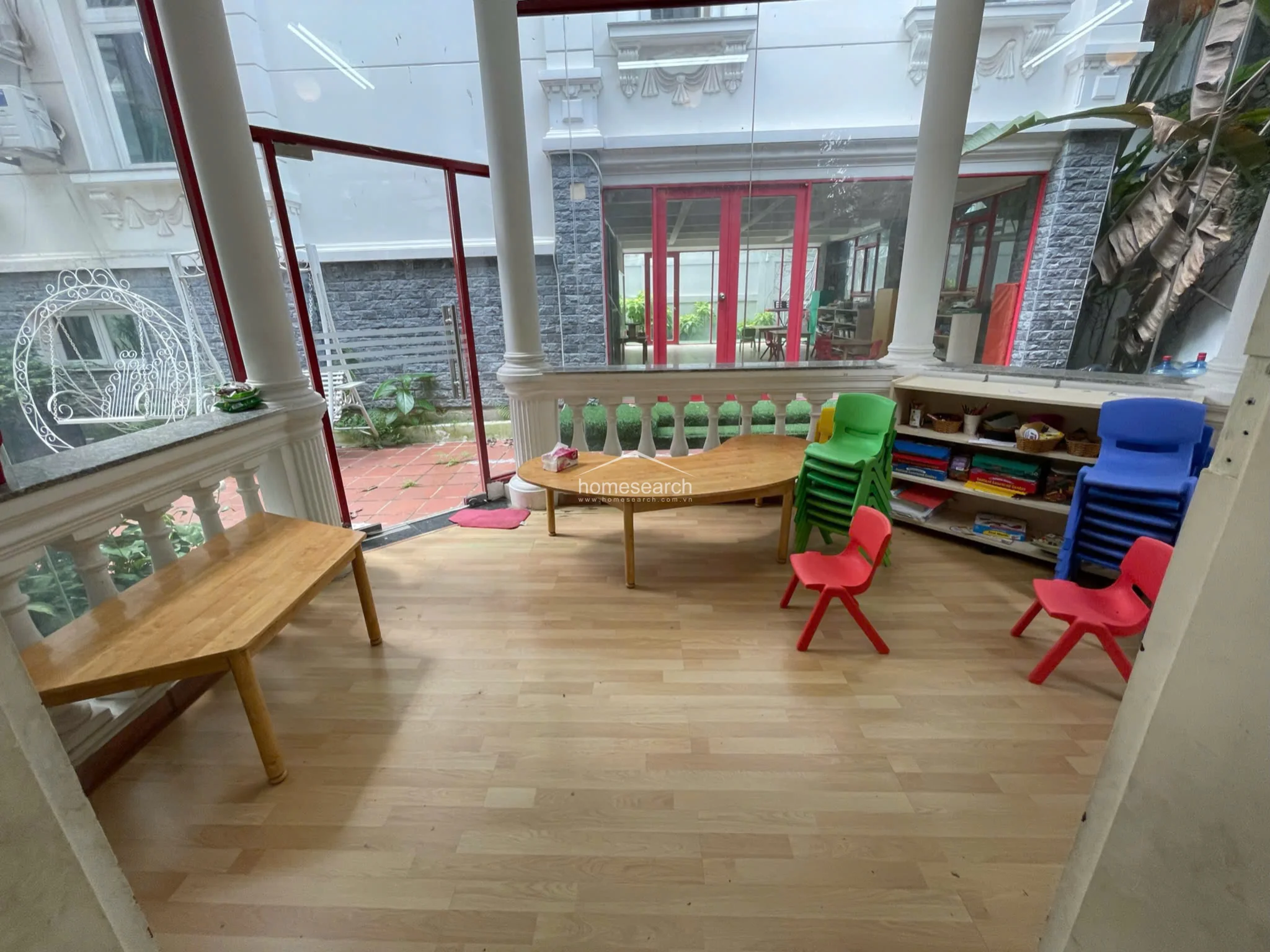 This sun-drenched room features light wood-look flooring and floor-to-ceiling glass walls that provide a seamless connection to an outdoor terrace area. The space is currently utilized as a children's activity room, furnished with low wooden tables, colorful plastic chairs, and open shelving units for storage. Classic architectural columns and a decorative balustrade add a touch of traditional elegance to the bright, open layout.