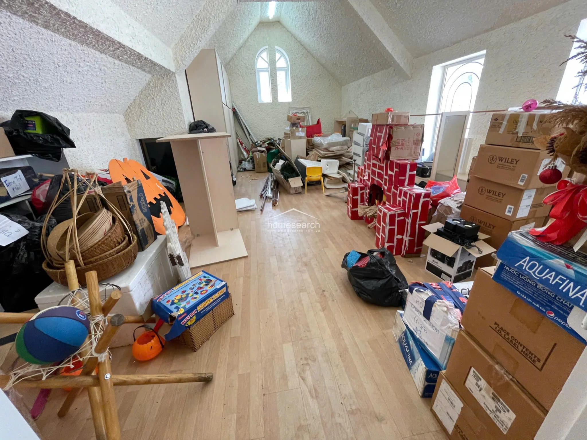This attic space is currently utilized as a storage area, filled with various boxes, hobby equipment, and miscellaneous items. It retains the same vaulted ceiling and light wood flooring as the adjacent room, with two arched windows providing natural light to the cluttered but structurally sound environment.