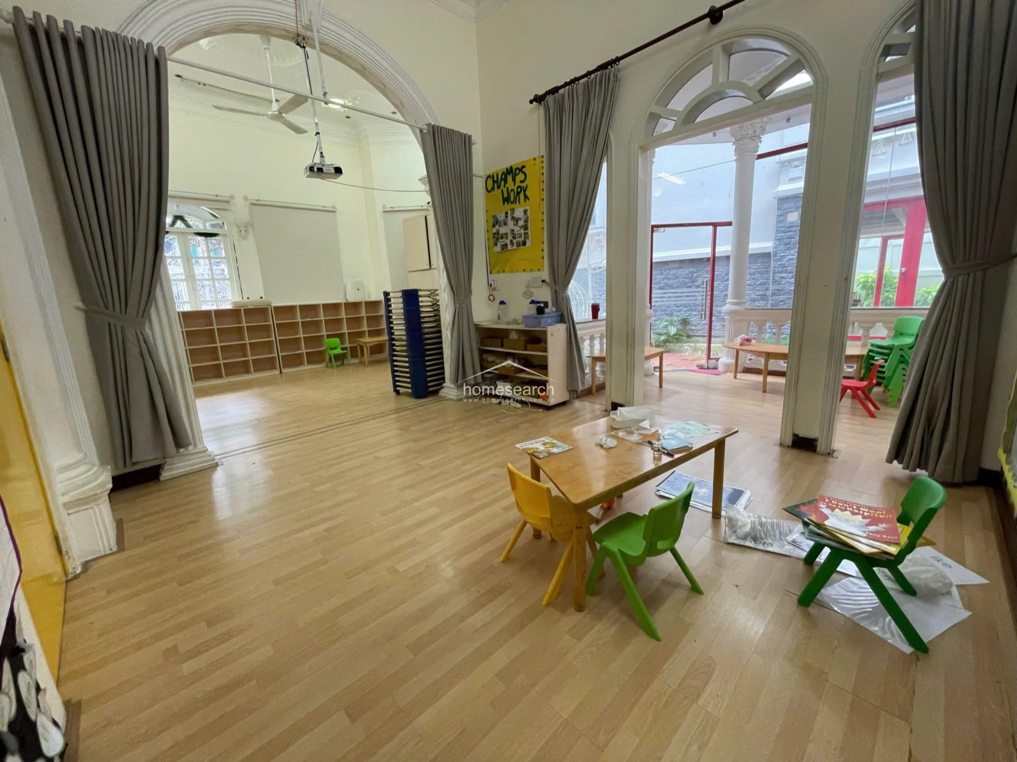 This spacious classroom area utilizes large arched openings and grey curtains to create a flexible, open-flow layout that connects to an outdoor terrace. The room is furnished with child-sized tables and chairs, features high ceilings with a mounted projector, and maintains a bright, airy feel with ample natural light.