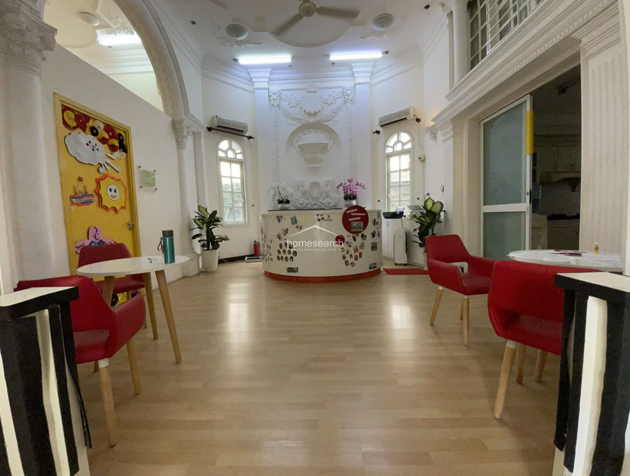 This grand reception area is defined by high ceilings with intricate molding, decorative wall reliefs, and elegant arched windows that allow for ample natural light. The space features a curved, central reception desk, light wood flooring, and a neutral color palette accented by vibrant red chairs. The room flows openly into adjacent areas through large archways, maintaining a sophisticated yet functional atmosphere.