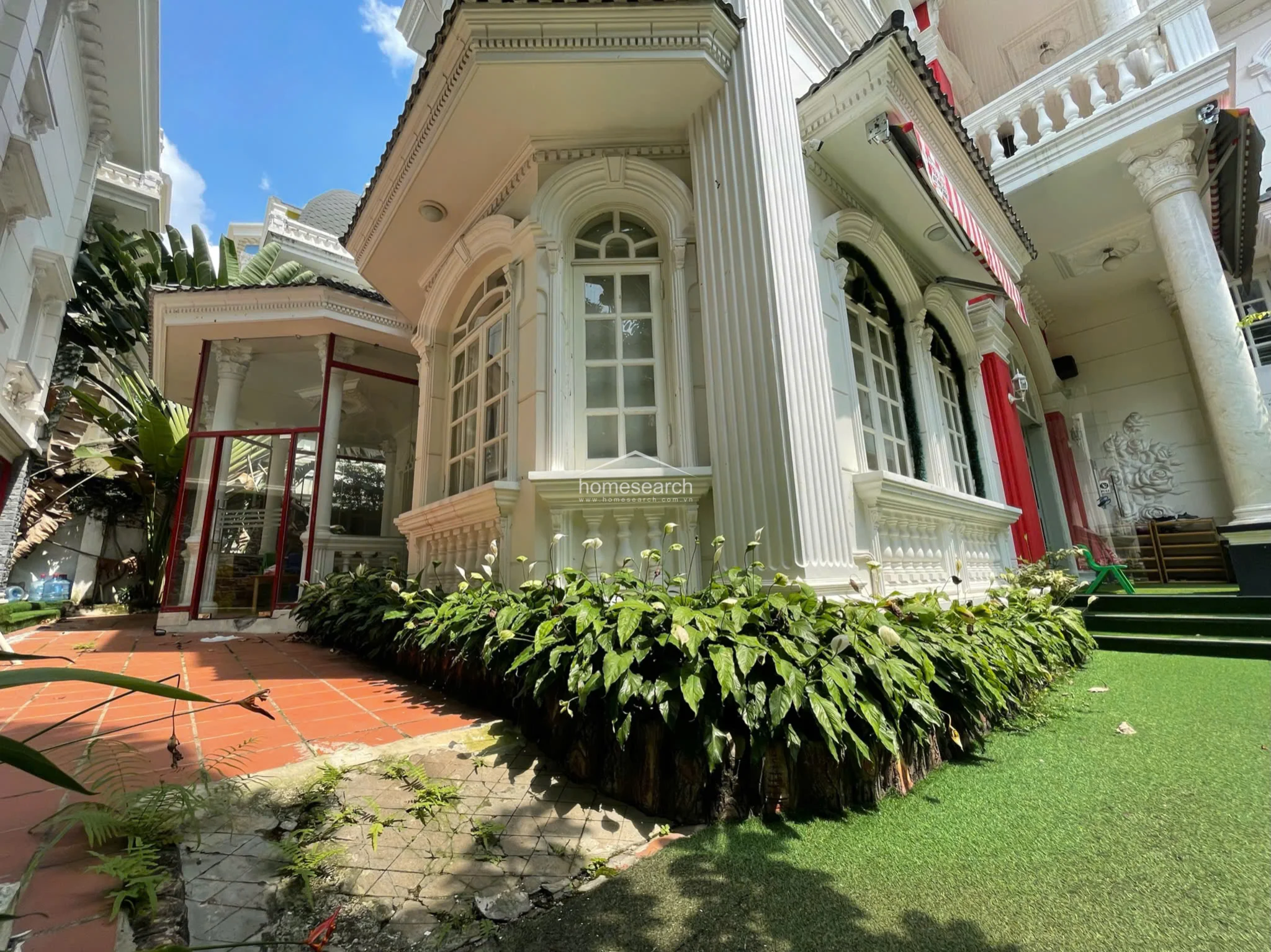 This close-up perspective emphasizes the elegant corner of the villa, featuring a glass-enclosed sunroom or conservatory area with red-framed windows. The landscaping is well-integrated, with vibrant peace lilies planted along the foundation, softening the formal lines of the neoclassical columns and white stucco walls.