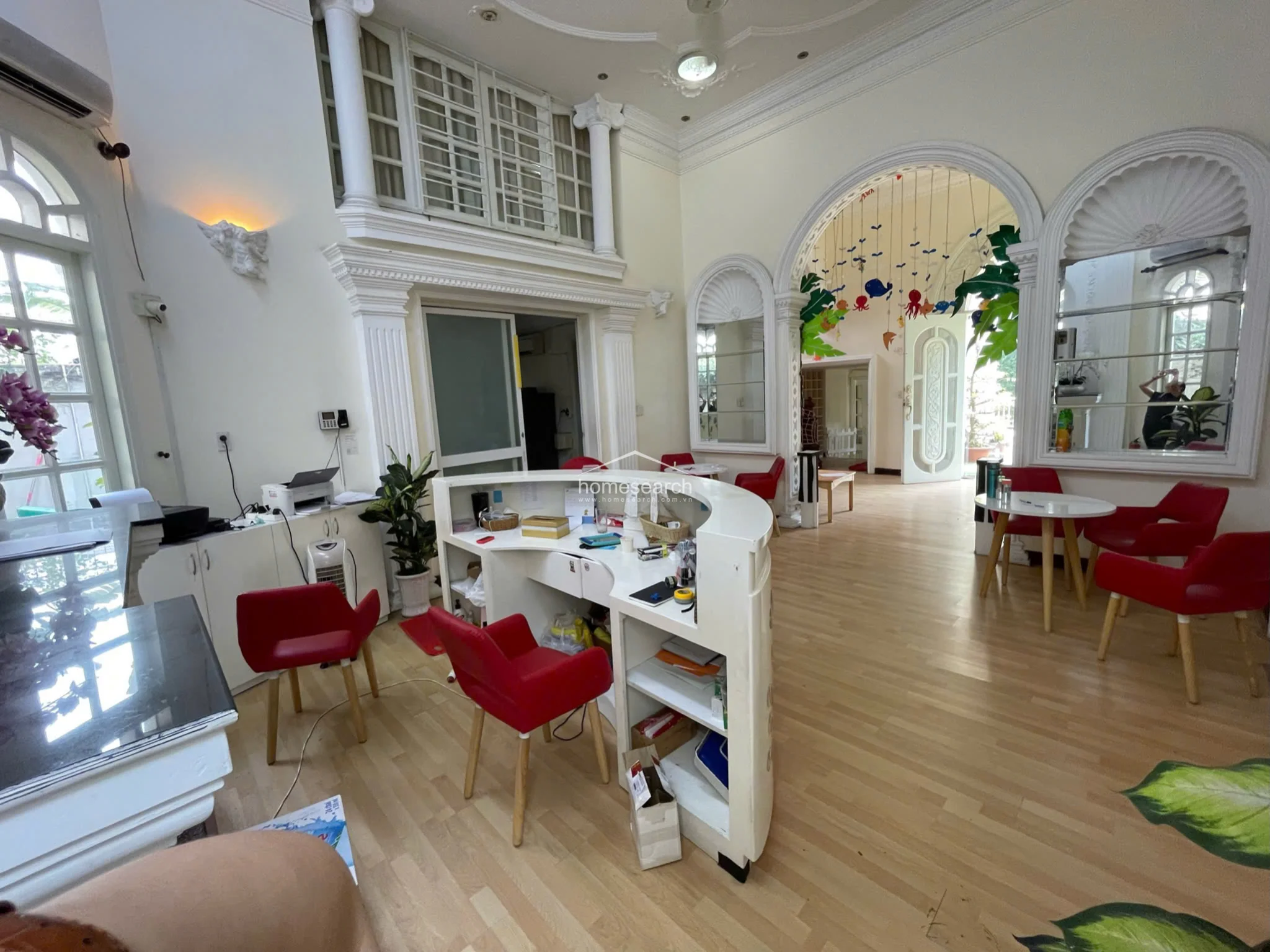 This reception area showcases impressive architectural details, including ornate ceiling medallions, classical columns, and arched wall niches. The space is bright and airy, connecting to other rooms via wide, open archways that create a fluid and inviting floor plan. It is well-furnished with a central desk and seating areas, all set against a clean, light-toned backdrop that emphasizes the room's height and character.