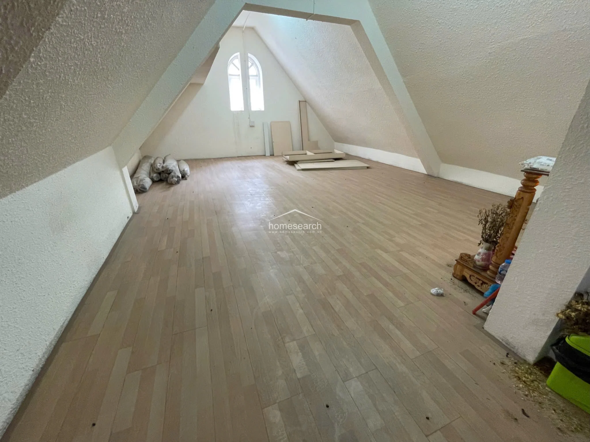 This attic-style room features a vaulted ceiling with textured white finishes and light wood flooring, providing a clean, minimalist aesthetic. The space is illuminated by an arched window at the far end and contains some loose construction materials, indicating it is currently an unfinished or flexible-use area.
