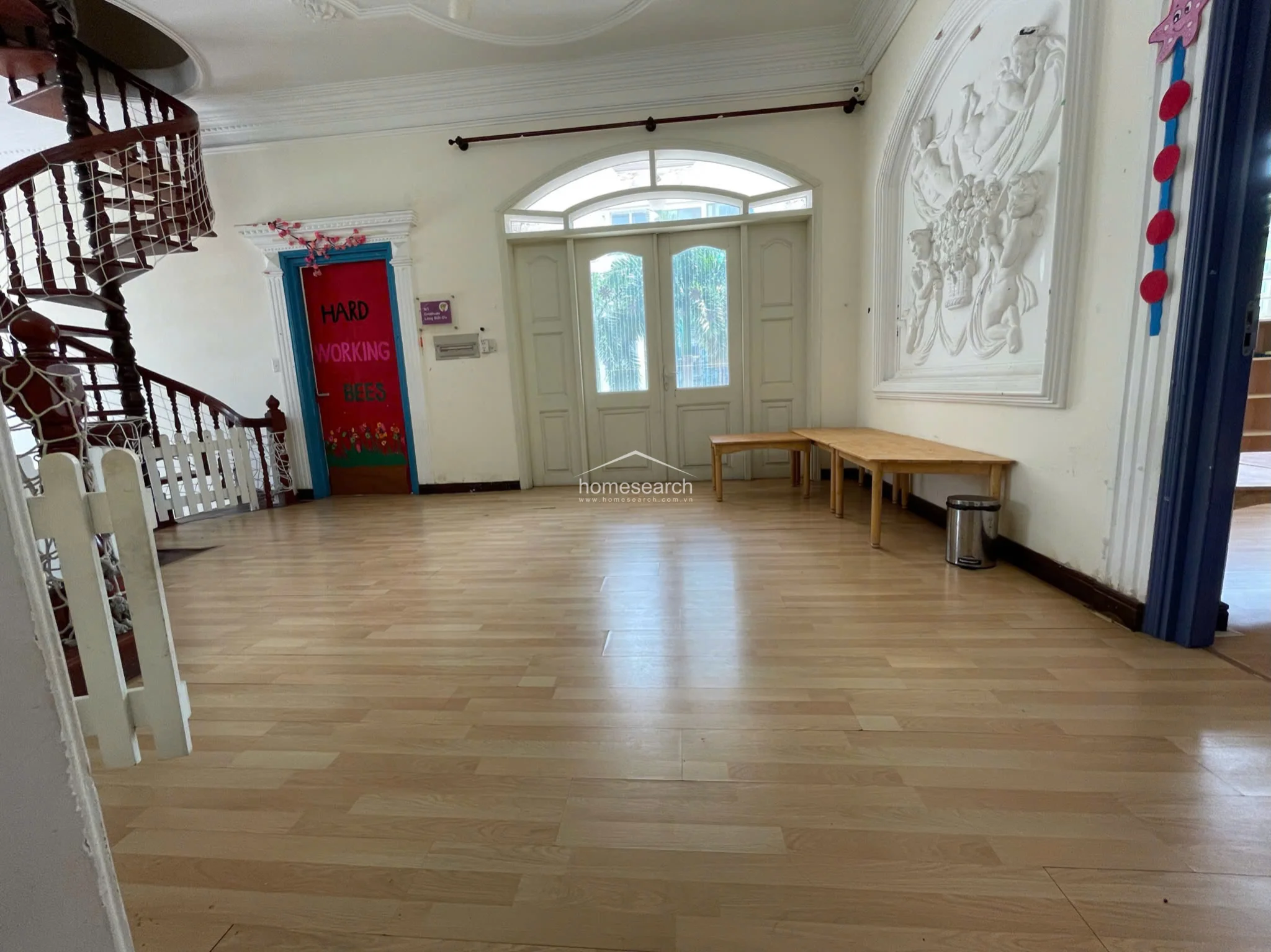 This spacious, open-plan room serves as a central hub, featuring light wood flooring and a striking, large-scale white relief sculpture on the wall. The space is defined by a grand spiral wooden staircase and an arched double-door entryway that allows for ample natural light, maintaining an airy and sophisticated atmosphere.