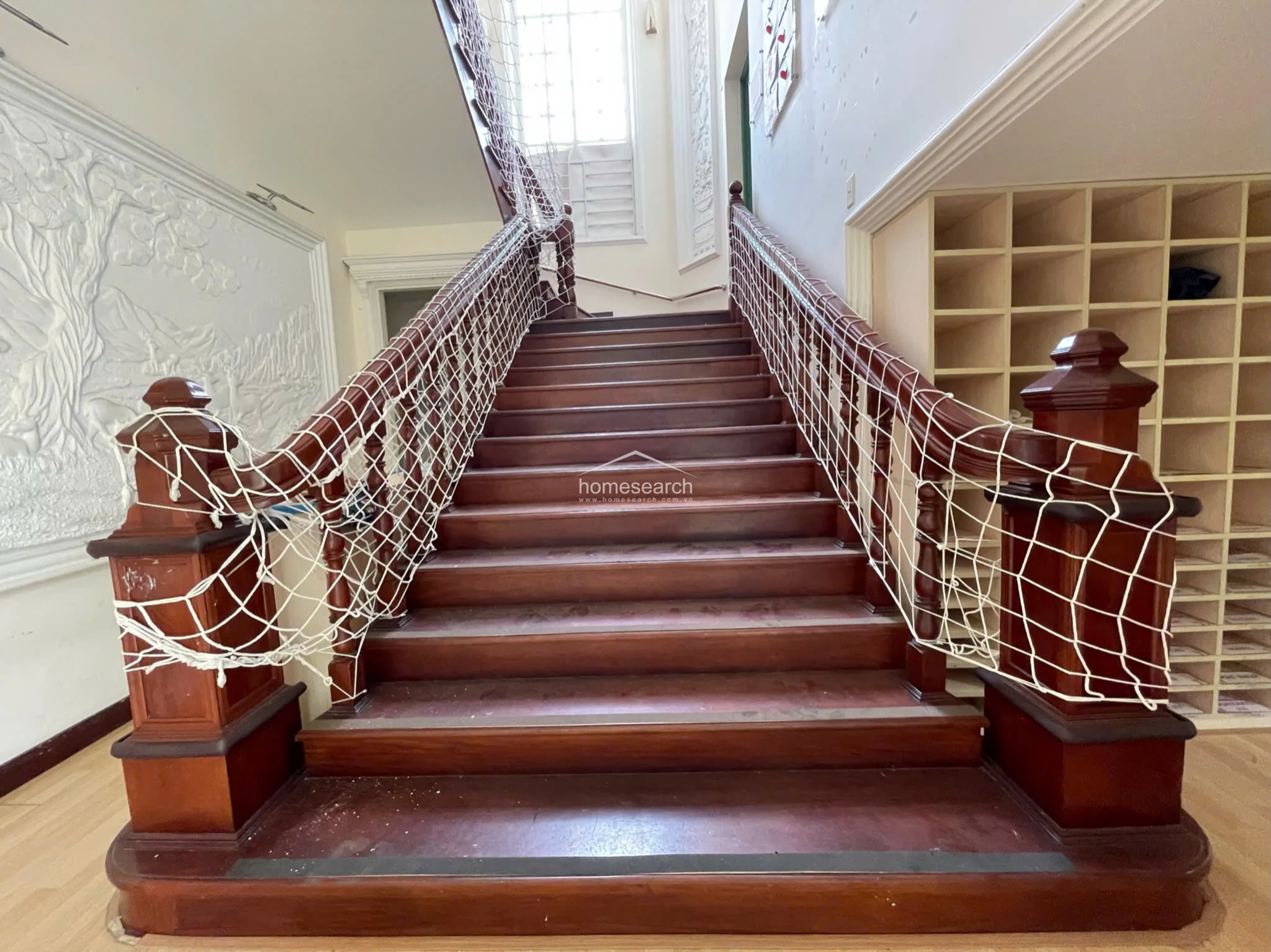 A close-up view of the staircase highlights the solid wood construction, decorative newel posts, and the safety netting installed for child protection. The surrounding walls feature elegant, classical-style relief molding, contrasting with the practical, modern cubby storage located to the right.