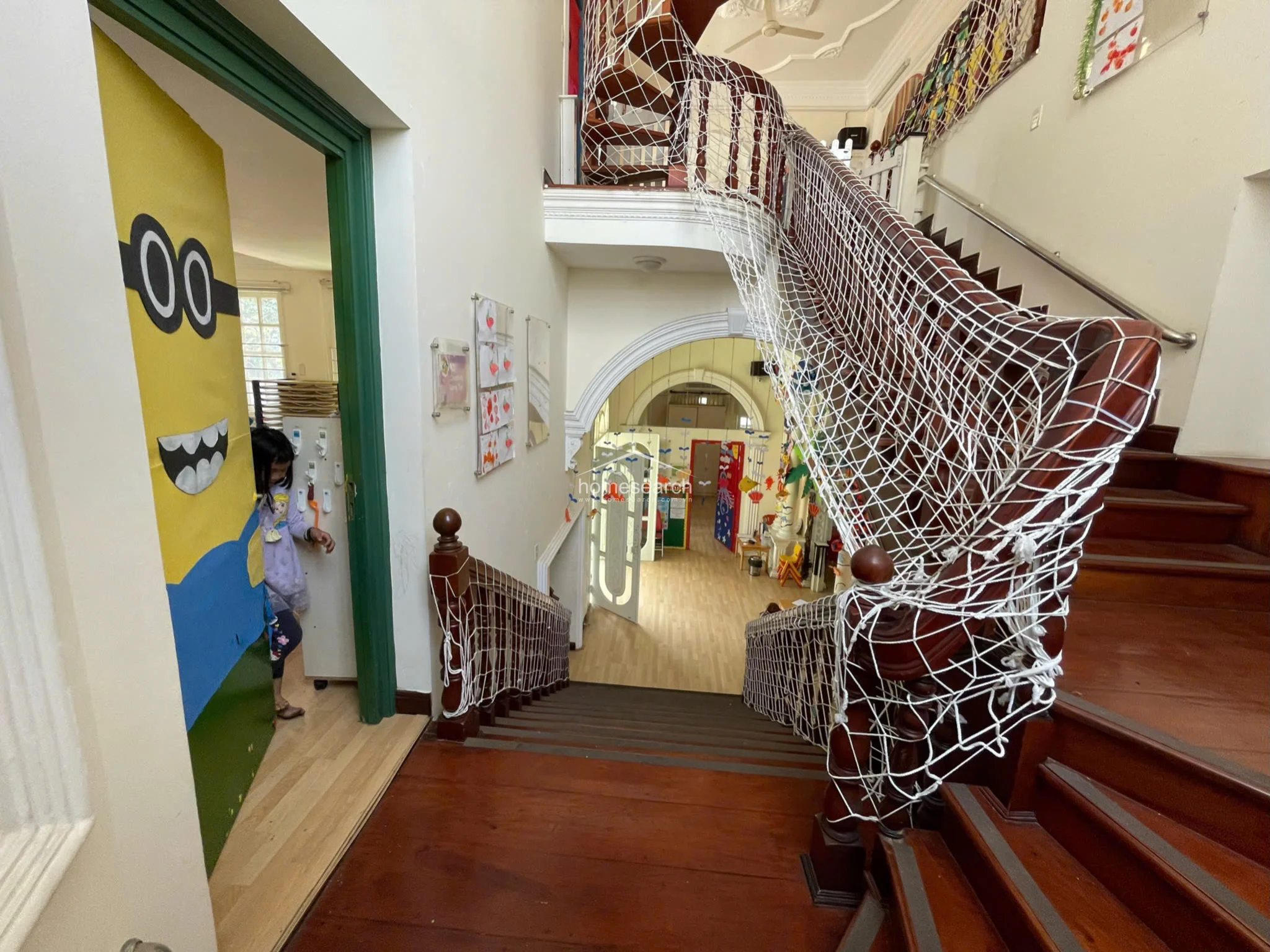 Looking down from the landing, this perspective emphasizes the elegant wooden staircase and the open-plan layout of the lower level. The space is brightly lit and connects directly to a room featuring a vibrant, character-themed door, highlighting the property's versatile and airy architectural flow.